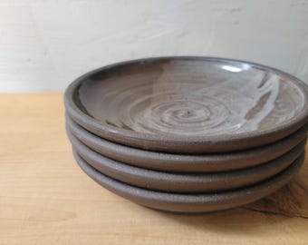 May include: A stack of four handmade ceramic plates. The plates are a dark brown color with a lighter brown swirl pattern in the center. The plates have a slightly rough texture and a simple, rustic design.