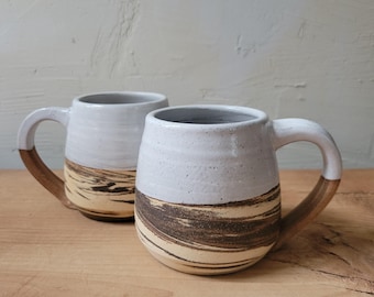 May include: Two ceramic mugs featuring a two-tone design with a smooth white glazed upper section and a lower section characterized by a swirling, marbled pattern of dark brown and tan earth tones, each equipped with a curved handle.