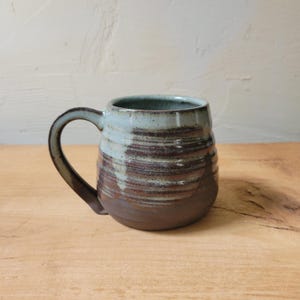 May include: A ceramic mug featuring a rounded body with a handle. The upper portion is glazed in a light blue-grey tone, while the lower section displays a dark brown, unglazed clay finish. Horizontal ridges texture the middle of the mug.