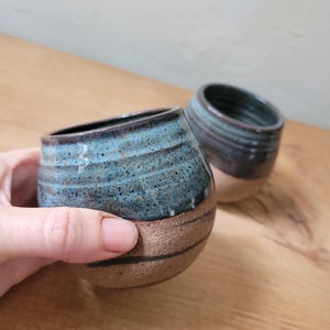May include: Two rounded ceramic cups featuring a speckled blue and dark teal glaze on the upper portion and a textured, unglazed tan clay base. The cups have horizontal ridges along the sides and a smooth, curved shape for holding.