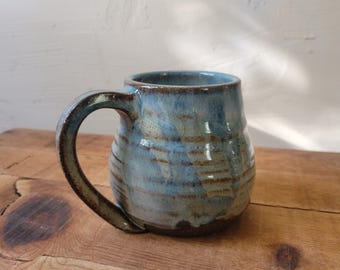 May include: A ceramic mug with a rounded body and a curved handle, featuring a variegated glaze in shades of blue and brown. The surface has horizontal ridges and a glossy finish, with a darker brown tone at the base and on the handle.