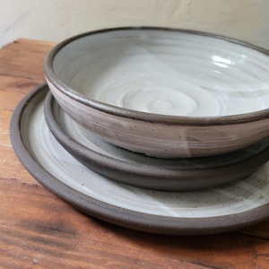 May include: A stack of three ceramic dishes consisting of a deep bowl and two flat plates. Each piece features a smooth, off-white interior with visible circular wheel-thrown markings and a contrasting dark brown, unglazed rim and exterior edge.