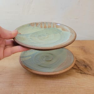 May include: Two round ceramic plates featuring a speckled tan clay base with a smooth, glossy glaze in shades of sage green and pale blue. The glaze has a swirled, mottled appearance with visible drips along the edges of the upper plate.