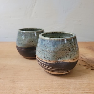 May include: Two rounded ceramic cups featuring a two-tone design. The upper portion of each cup has a speckled light blue and green glaze, while the lower portion displays a dark brown, unglazed clay base with visible horizontal texture lines.