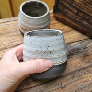 May include: Two ceramic cups with a rounded shape and a two-tone finish. The upper portion features a speckled off-white glaze with horizontal ridges, while the lower portion is a smooth, solid dark charcoal gray. The cups have an open top.