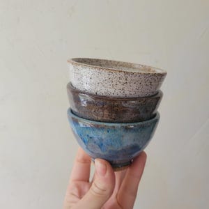 May include: A stack of three small ceramic bowls. The top bowl is off-white with dark brown speckles. The middle bowl is a solid dark brown with a glossy finish. The bottom bowl features a blend of deep blue and brown tones with a smooth glaze.