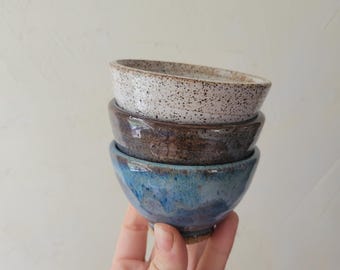 May include: A stack of three small ceramic bowls. The top bowl is off-white with dark brown speckles. The middle bowl is a solid dark brown with a glossy finish. The bottom bowl features a blend of deep blue and brown tones with a smooth glaze.