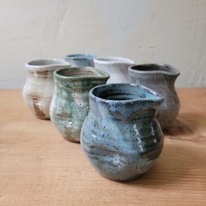 May include: A collection of five small ceramic pitchers with varied glaze finishes including blue, green, white, and grey. Each pitcher features a rounded body, a small pouring spout, and a textured, hand-thrown appearance with unique color variations.