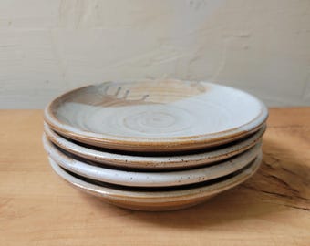 May include: A stack of four round ceramic plates with a speckled white glaze on the interior and a natural tan clay rim. The plates feature a shallow basin and a slightly raised edge, showcasing a hand-thrown appearance with visible circular textures.