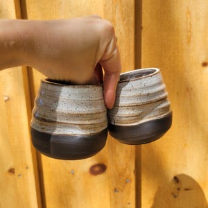 May include: Two ceramic cups featuring a speckled off-white upper section with horizontal ridges and a smooth, solid dark brown base. The cups have a rounded, bulbous shape and a wide opening at the top, showcasing a contrasting two-tone design.
