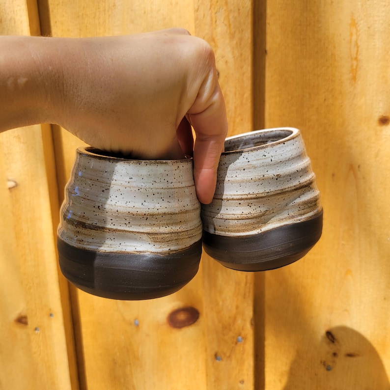 May include: Two ceramic cups featuring a speckled off-white upper section with horizontal ridges and a smooth, solid dark brown base. The cups have a rounded, bulbous shape and a wide opening at the top, showcasing a contrasting two-tone design.