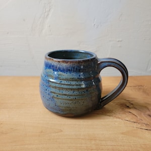May include: A handmade ceramic mug with a rounded body and a large handle. The mug features a blue glaze with brown and tan accents, creating a textured, rustic appearance. The mug is sitting on a wooden surface.