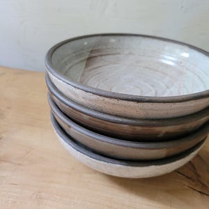 May include: A stack of four ceramic bowls with a speckled off-white interior and a dark brown exterior. The bowls feature a visible circular texture on the inner base and a smooth, rounded rim. The glaze shows subtle variations in color and tone.
