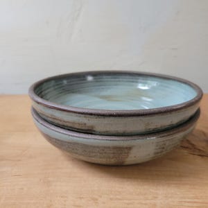 May include: Two stacked ceramic bowls featuring a light blue glazed interior and a dark brown, textured exterior rim. The bowls have a circular shape with visible horizontal ridges along the inner walls and a smooth, rounded edge at the top.