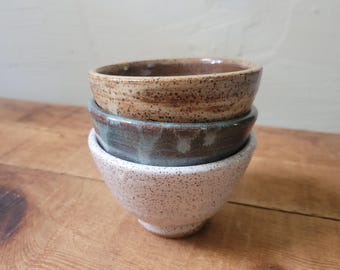 May include: A stack of three small ceramic bowls. The top bowl is tan with dark brown speckles. The middle bowl features a smooth, dark blue-grey glaze. The bottom bowl is light pink with fine dark speckles, featuring a tapered base design.