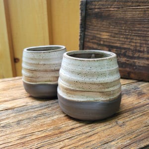 May include: Two ceramic cups with a rounded, ribbed shape. The upper portion of each cup features a speckled off-white glaze, while the lower portion is finished in a solid dark brown tone. The cups have a wide opening and a smooth, curved base.