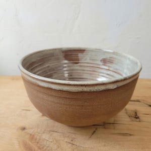 May include: A brown ceramic bowl with a white rim and a white glaze inside. The bowl has a textured surface and is slightly wider at the top than the bottom.