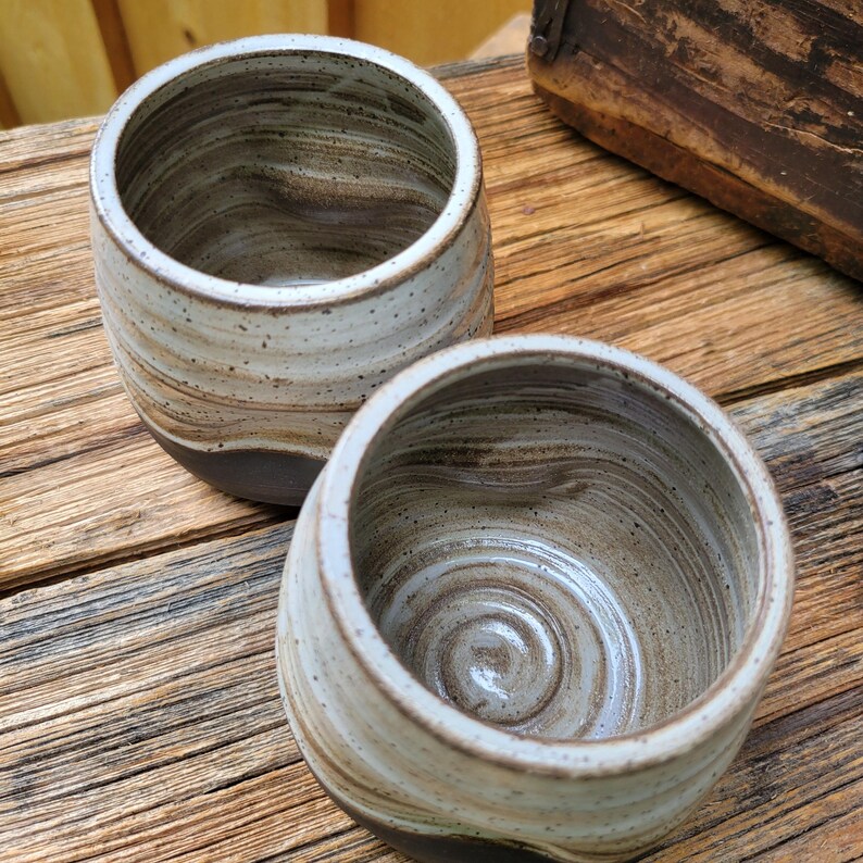 May include: Two ceramic cups featuring a marbled pattern of cream, beige, and dark brown tones. The exterior and interior surfaces display a smooth, glossy finish with subtle speckles throughout the clay, showcasing a rounded, handleless design.
