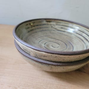 May include: A stack of two round ceramic bowls featuring a speckled cream-colored glaze with visible dark brown spiral patterns on the interior. The bowls have a thick, dark brown rim and a textured, earthy finish on the exterior surface.