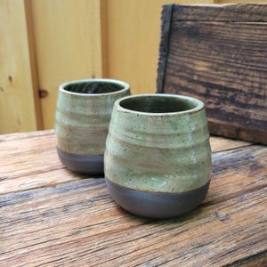 May include: Two handmade ceramic tumblers. The cups have a rounded shape with a dark brown base and a light green and tan upper section. The cups are sitting on a wooden surface.
