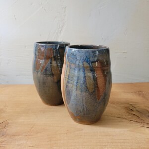 May include: Two cylindrical ceramic tumblers featuring a reactive glaze with overlapping shades of deep blue, earthy brown, and muted gray. The surface displays a smooth, glossy finish with vertical drip patterns and subtle color variations throughout.
