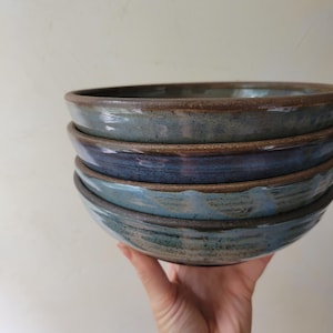 May include: A stack of four round ceramic bowls with a dark brown rim and a mottled glaze featuring shades of blue, grey, and earthy brown. The bowls have a smooth, slightly reflective surface and a visible texture along the exterior sides.