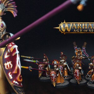 May include: A group of miniature figures painted in gold, white, and red, holding spears and shields. The figures are wearing elaborate helmets and armor. The figures are standing on a black background. The text "Warhammer Age of Sigmar" is visible in the background.