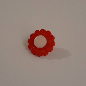 May include: A red flower-shaped button with a pale pink centre. The button has a scalloped edge and is set against a white background. The button is a small, decorative item.