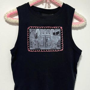 May include: Black sleeveless top with a crew neck. The top features a rectangular patch with a carousel design in shades of gray, framed with pink stitching. The top is hanging on a white and pink crocheted hanger.