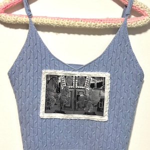 May include: A light blue cable knit crop top with spaghetti straps. The top features a black and white image of a carousel with horses, framed by a white border. The top is hanging on a crochet hanger.