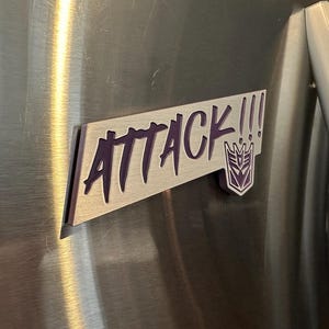 May include: A rectangular magnet with the word "ATTACK!!!" in purple lettering. The magnet also features a purple emblem. The magnet is attached to a stainless steel surface.