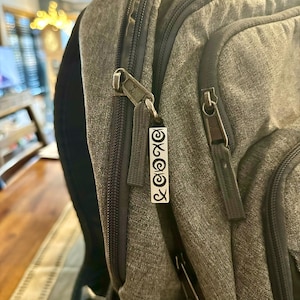 Puede incluir: Una mochila gris con un tirador de cremallera que presenta un amuleto plateado con el texto "XOXO" en él.