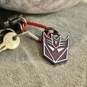 May include: A purple and silver Transformers keyring with a red carabiner clip attached to a set of keys. The keyring features the Autobot symbol.
