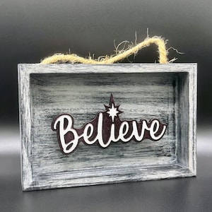 May include: A rustic wooden sign with a distressed gray finish. The sign features the word "Believe" in a burgundy color with a white star above it. The sign has a twine loop for hanging.