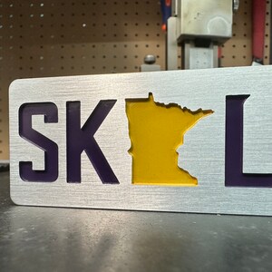 Può includere: Un cartello di metallo con le lettere "SK" e "L" in viola e il contorno dello stato del Minnesota in giallo.