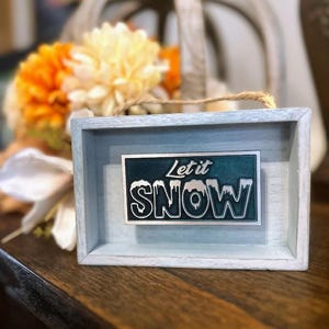 May include: A white wooden frame with a blue and white sign that says "Let it Snow". The sign has a snowflake design and is on a wooden table.