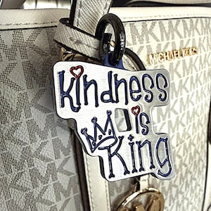 May include: A silver-colored keychain with the words "Kindness is King" in blue lettering, a red heart, and a crown design. The keychain is attached to a white and beige handbag with the "Michael Kors" logo.