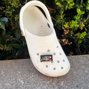 May include: A white Crocs clog with a black and colorful M&M's shoe charm. The charm features the M&M's logo and candy pieces. The shoe is on a concrete surface with green foliage in the background.