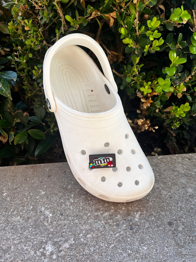 May include: A white Crocs clog with a black and colorful M&M's shoe charm. The charm features the M&M's logo and candy pieces. The shoe is on a concrete surface with green foliage in the background.