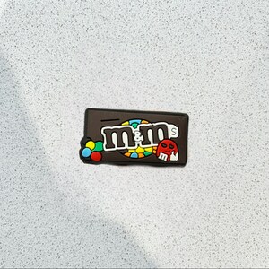 May include: A brown rectangular charm featuring the M&M's logo in white, with a colorful candy design. A small red skull is on the right side. The charm is likely made of rubber or a similar material.