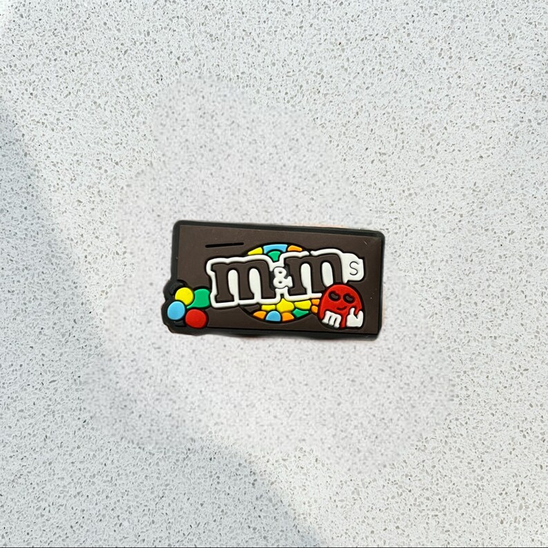 May include: A brown rectangular charm featuring the M&M's logo in white, with a colorful candy design. A small red skull is on the right side. The charm is likely made of rubber or a similar material.