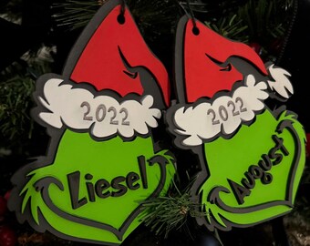 Puede incluir: Dos adornos navideños verdes y rojos con temática de Grinch con el año 2022 y los nombres "Diesel" y "August" escritos en ellos.