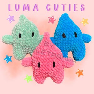 Puede incluir: Tres juguetes de peluche con forma de estrella en tonos turquesa, rosa y azul, cada uno con ojos negros y pequeños brazos. Están sobre un fondo rosa con estrellas y el texto "LUMA CUTIES".