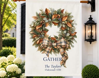 Könnte beinhalten: Eine rechteckige, cremefarbene Gartenflagge mit einem herbstlichen Kranz aus grünen und orangefarbenen Blättern sowie einer großen karierten Schleife. Darunter steht in dunkelblauer Schrift GATHER, The Taylors und Oakwood, OH.