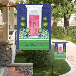 May include: A navy blue garden flag with a watercolor design. The flag features a pink door, green topiary, and blue and white vases. The text "The Stewarts" is displayed on a green and pink stripe.