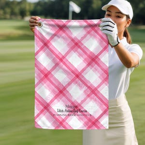 May include: A pink and white plaid golf towel with the text "Maggie, Witch Hollow Golf Course, PUMPKING RIDGE GOLF CLUB". The towel is held up by a person wearing a white golf glove and a white hat. The background is a golf course.