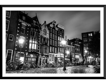 May include: A black and white photograph of a street scene in Amsterdam at night. The street is lined with buildings, and there are streetlights and bicycles parked along the side of the road.