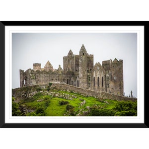 May include: A gray stone castle with multiple towers and a tall, pointed roof sits on a grassy hill. The castle is surrounded by a stone wall and there are trees in the foreground.