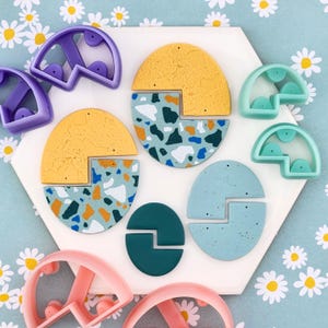 May include: A collection of colorful polymer clay earrings and matching cutters. The earrings feature a terrazzo design with yellow, blue, and teal accents. The cutters are in shades of pink, purple, and teal. The earrings are arranged on a white hexagonal tile.