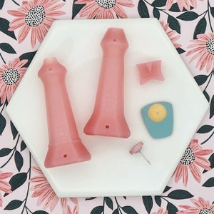 May include: A set of five pink silicone molds for making candles. The molds are shaped like a tall, slender tower, a small, flat rectangle, a small, round disc, and a small, pointed cone.